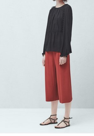 Blusa nera a maniche lunghe con design a pieghe, culotte rosse a gamba larga e sandali neri con borchie. Tessuto morbido con dettagli testurizzati.