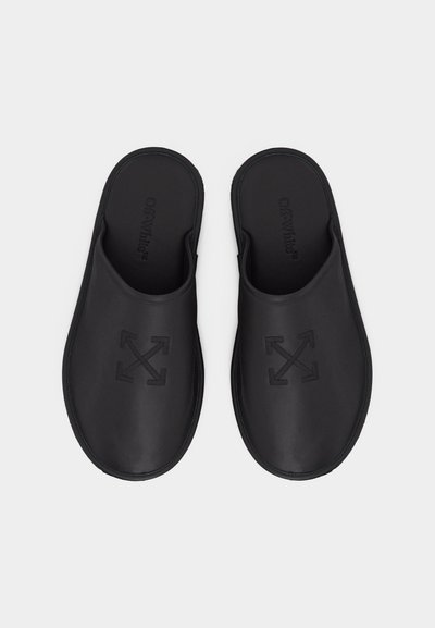 Μαύρα παπούτσια τύπου slip-on με κλειστές μύτες και ανάγλυφο σχέδιο με διασταυρούμενα βέλη στην επάνω επιφάνεια, επίπεδη σόλα και ανοιχτή πίσω πλευρά.