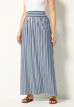 Femme portant une jupe longue taille haute à rayures verticales bleu marine et blanc et des sandales ouvertes marron, debout devant un fond blanc.