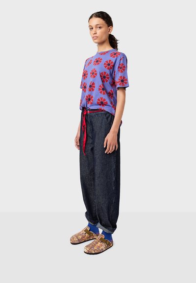 Camiseta morada con estampados de flores rojas, combinada con pantalones sueltos de rayas verticales negras y zapatillas multicolores de tipo slip-on con hebilla.
