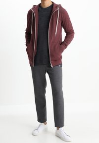 Bordeauxfarbene Zip-up-Hoodie mit weißen Kordeln, kombiniert mit einem strukturierten dunkelgrauen Pullover und grauen Hosen. Weiße Sneakers runden das Outfit ab.