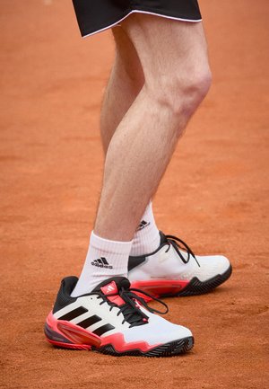 Hvide og sorte atletiksko med røde accenter, teksturerede såler, markante sorte striber og sorte snørebånd, kombineret med hvide crew-sokker.