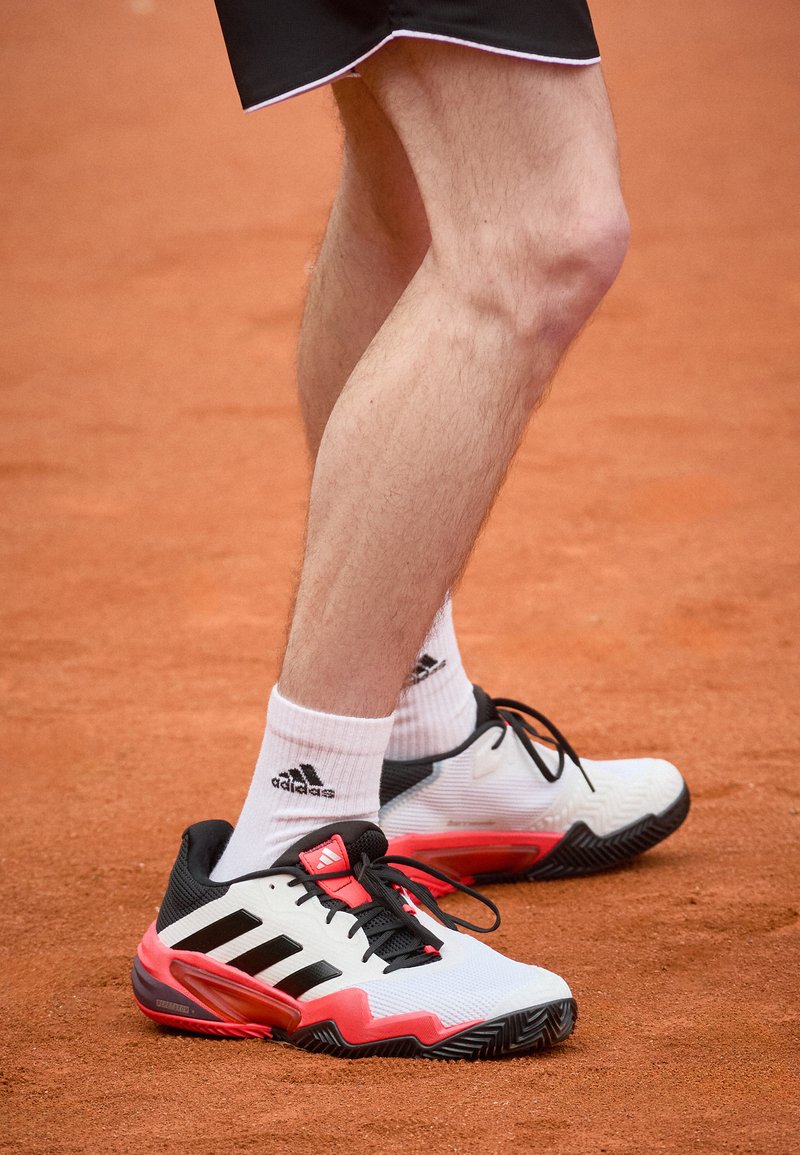 Vita och svarta sportskor med röda detaljer, texturerade sulor, framträdande svarta ränder och svarta snören, kombinerade med vita mellanhöga strumpor.