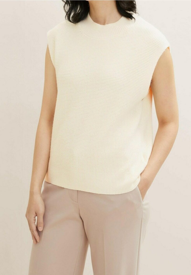 Femme portant un haut en maille crème sans manches et un pantalon beige, une main dans la poche, sur un fond beige clair.
