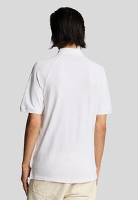 Hvid polo-shirt med korte raglanærmer, glat tekstur og en klassisk krave. Vist fra bagview, med et enkelt design.