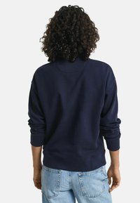 Persoon met krullend haar, gekleed in een navyblauwe trui en lichtblauwe spijkerbroek, staat met de rug naar voren tegen een effen achtergrond.