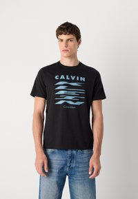 Juodas medvilnės marškinėliai su mėlyna grafine užrašu "CALVIN" ir bangomis. Apvalus iškirpimas ir trumpi rankoviai. Derinami su šviesiai mėlynomis džinsomis.