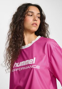 Rosa idrottströja med vita detaljer, som har en ribbad textur och logotypen "hummel PERFORMANCE" tydligt synlig i vitt.