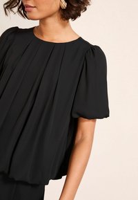 Fekete plisezett blúz rövid, puffos ujjal és kerek nyakkal. Puha, folyékony anyag vízszintes plissékkel, amelyek laza sziluettet alakítanak ki.
