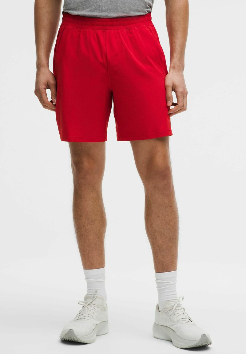 Homme portant un short de sport rouge, des chaussettes blanches montantes et des chaussures de course blanches, debout devant un fond clair uni.