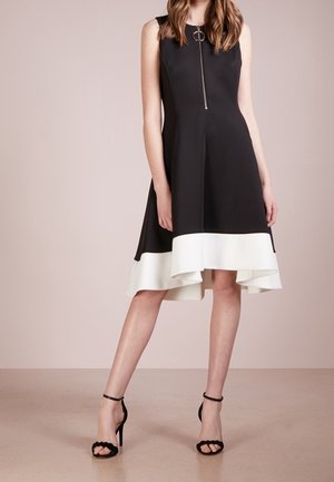 Robe noire sans manches avec fermeture éclair à l'avant et ourlet asymétrique blanc, portée avec des sandales noires à talons hauts et brides à la cheville.