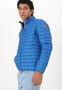 Blauwe pufferjack met horizontaal geweven ontwerp, ritssluiting aan de voorkant, hoge kraag en een oranje ritssluiting. Bevat een circulair logo op de mouw.
