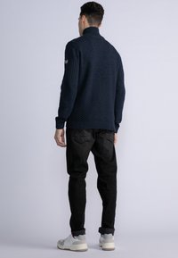 Mörkblå stickad tröja med hög krage och ribbad textur, kombinerad med svarta byxor och grå-vita sneakers. Har en logopatch på sidan.