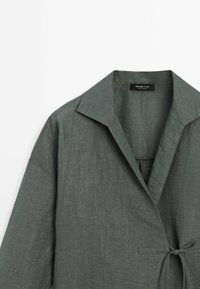 Chemise légère en lin vert foncé avec col, boutonnage à l'avant et fermeture par lien. Tissu texturé et coupe décontractée.