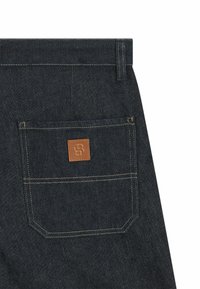 I pantaloni in denim blu scuro presentano una tasca posteriore cucita con un'etichetta rettangolare in pelle marrone. La texture è liscia con una leggera lucentezza.