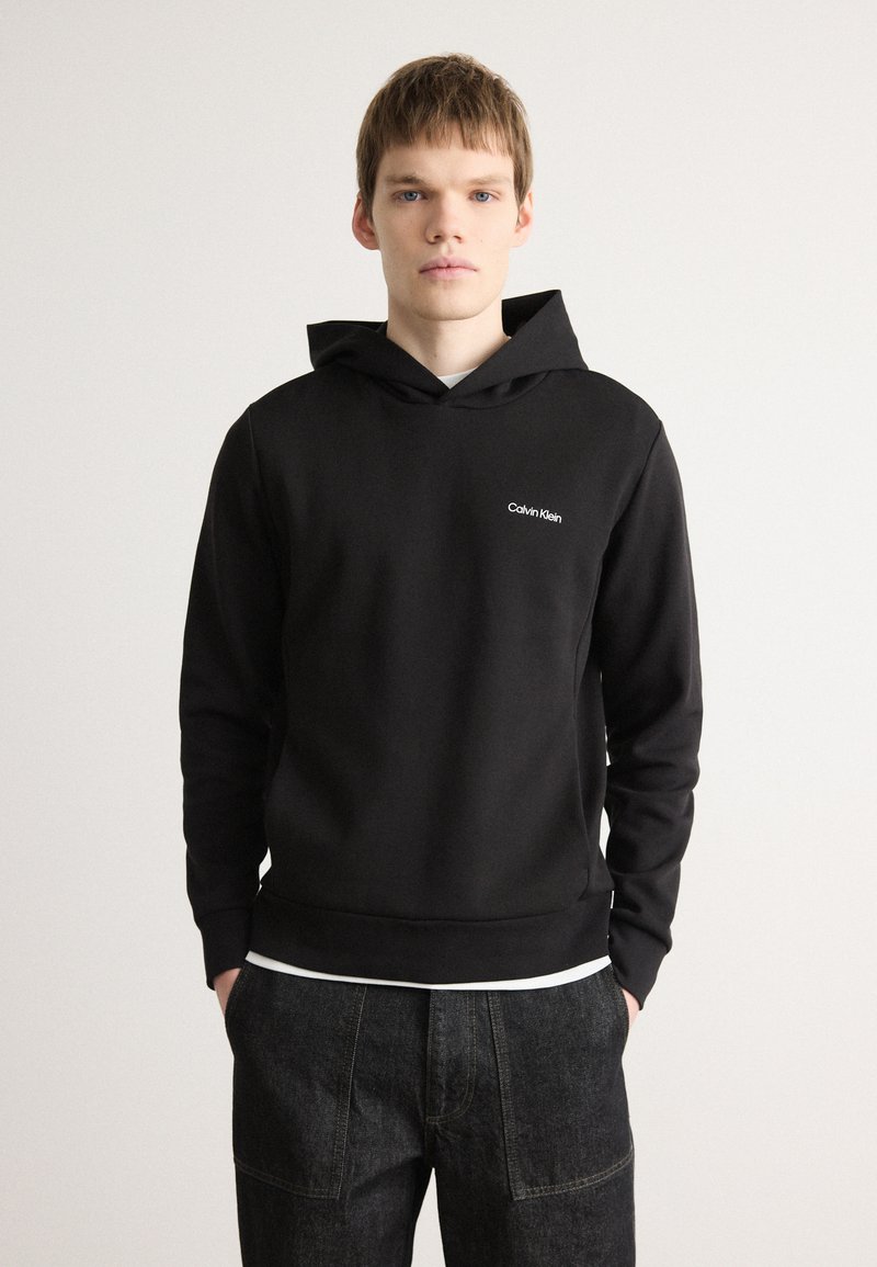 Svart hettegenser med lommer foran, laget av myk stoff, har en liten hvit "Calvin Klein" logo på brystet.