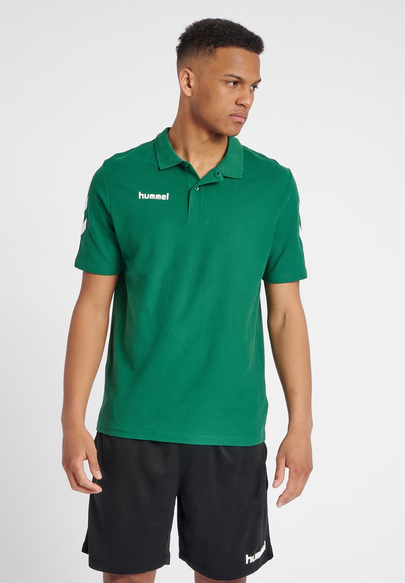 Grøn polo med krave, korte ærmer og Hummel-logo. Har hvide chevrons på ærmerne og en glat tekstur. Bæres med sorte shorts.