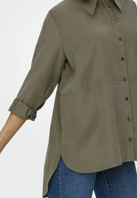 Camicia a maniche lunghe verde oliva con colletto, frontale con bottoni e polsini arrotolati. Realizzata in materiale morbido con una vestibilità leggermente ampia e spacchi laterali.