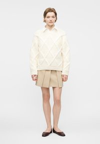 Νεαρή γυναίκα που φοράει ένα φαρδύ εκρού πουλόβερ με πλεκτό σχέδιο κοτσίδας, μπεζ μίνι φούστα με πιέτες και σκούρα καφέ μπαλαρίνες, στέκεται μπροστά σε ένα απλό φόντο.