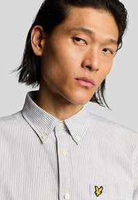 Camicia a maniche lunghe a righe bianca e grigia con colletto a punta, caratterizzata da un piccolo logo giallo ricamato sul taschino a sinistra.