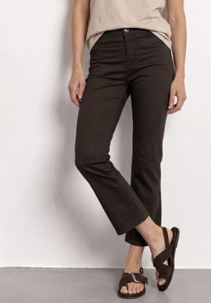 Frau trägt dunkelbraune Slim-Fit-Hose und braune Wildleder-Sandalen mit Schnallenriemen, steht vor einer weiß strukturierten Wand.