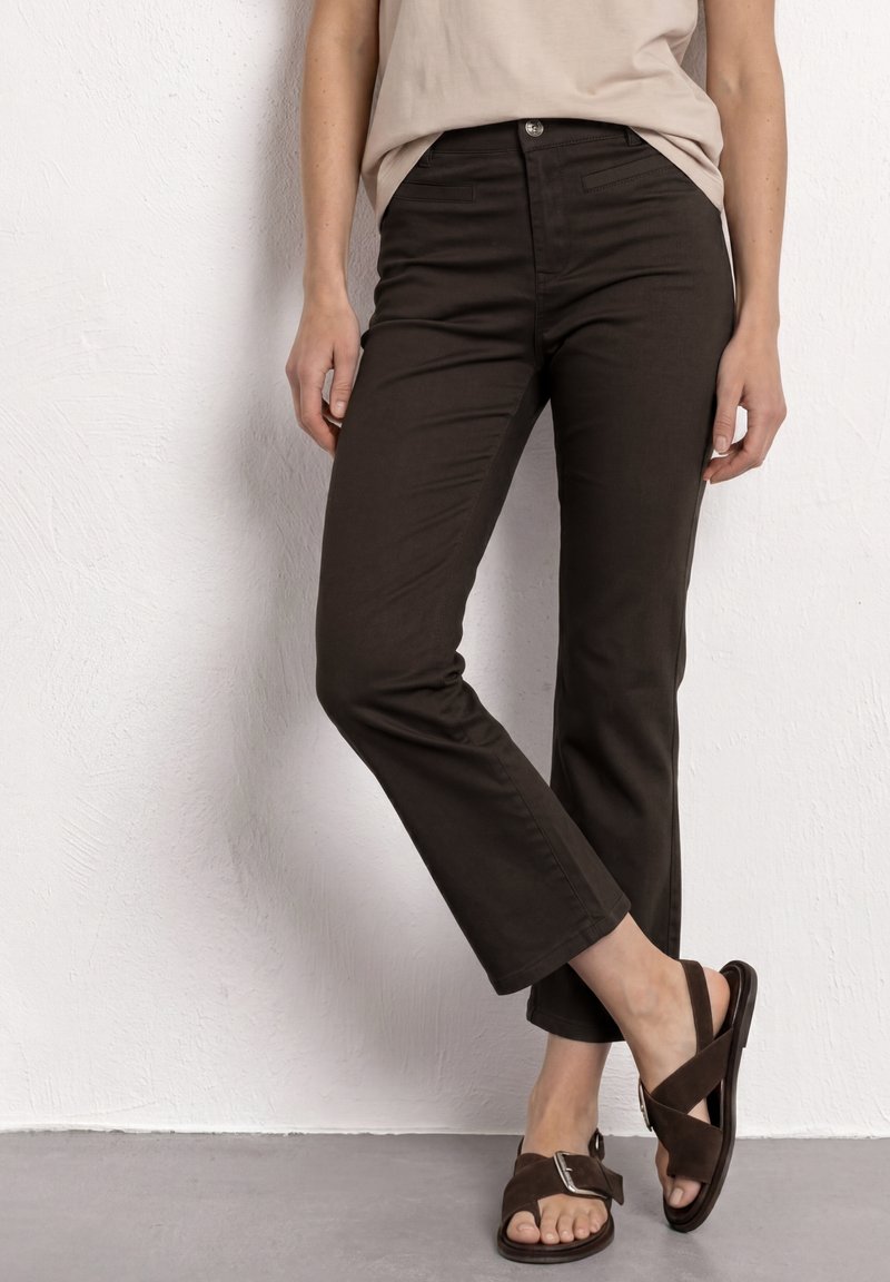 Vrouw draagt donkerbruine slim-fit broek en bruine suède sandalen met gespen, staand tegen een witte getextureerde muur.