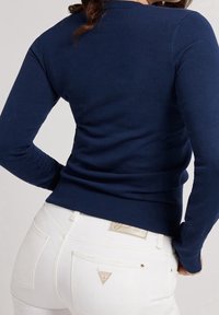 Marineblauer, figurnaher Pullover mit langen Ärmeln, gerippten Bündchen und Saum. Kombiniert mit weißen Jeans, die ein Logo-Patch und Gesäßtaschen haben.