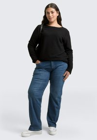 Schwarzes, strukturiertes Langarmoberteil, kombiniert mit hellblauen Jeans in gerader Form und weißen Sneakers. Das Model steht mit einer Hand in der Hüfte.