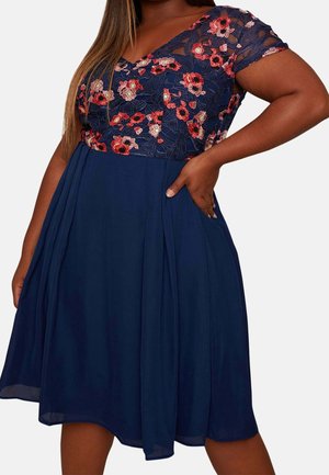 Mujer con un vestido azul marino con corpiño de encaje bordado con flores y mangas cortas, mano apoyada en la cadera.