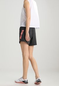 Osoba v bílém bezramenném topu, tmavě šedých šortkách s nápisem "JUST DO IT." a bílých teniskách Nike, stojící na jednobarevném pozadí.