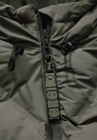 Giacca imbottita verde oliva con colletto alto, dotata di zip in plastica e cordini regolabili. Logo distintivo sul cursore della zip.