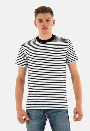 T-shirt a righe bianche e nere a maniche corte con colletto girocollo in maglia nera e piccolo logo sul lato sinistro del petto. Indossato con jeans blu.