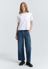 Tricou alb din bumbac cu mâneci scurte, combinat cu blugi evazați albaștri din denim și ghete negre până la gleznă, creând o ținută casual.