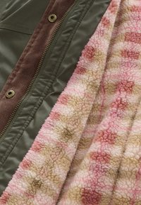 Zöld dzseki barna kordbársony szegéllyel, fém patentgombokkal, félig nyitott cipzárral, amely rózsaszín és bézs virágos polár bélést mutat.