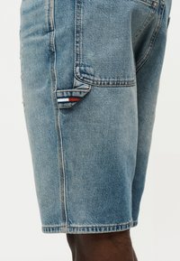 Ανοιχτό μπλε τζιν σορτς με πλαϊνή τσέπη που διαθέτει μια μικρή υφασμάτινη λεπτομέρεια με κόκκινες, λευκές και μπλε ρίγες σε ένα άτομο με σκούρο δέρμα.