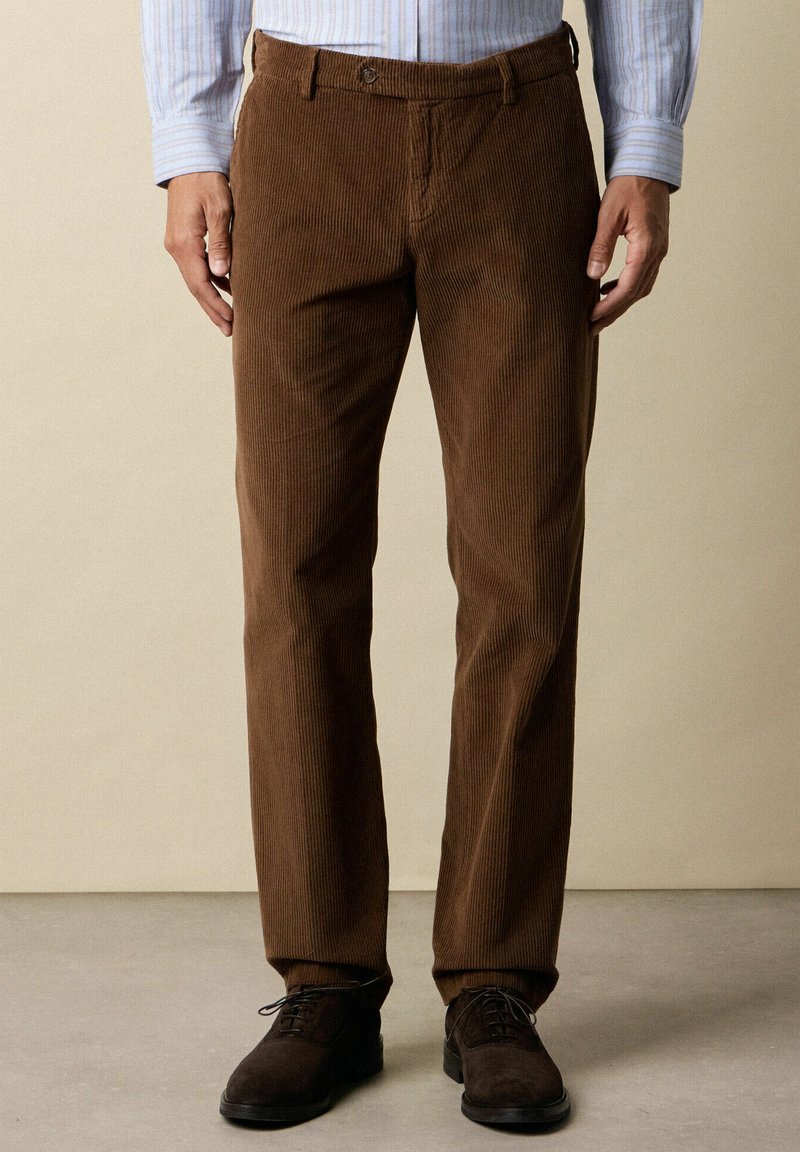 Pantaloni di velluto a coste marroni con design a gamba dritta, caratterizzati da righe verticali e tasche laterali, abbinati a scarpe scure e strutturate.