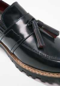 Mocassin en cuir noir avec glands, coutures détaillées et semelle en caoutchouc épaisse avec une bande de roulement prononcée, présenté en gros plan.