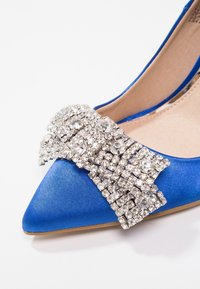 Chaussure plate en satin bleu à bout pointu avec un grand nœud de strass étincelants sur le devant et une doublure intérieure beige.