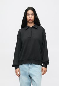 Mulher com cabelo longo e preto, vestindo um suéter solto preto com colarinho e jeans claros em um fundo branco liso.