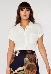 White, short-sleeve button-up shirt with a pocket. Textured fabric. Worn with a floral-patterned navy skirt featuring orange and green accents.