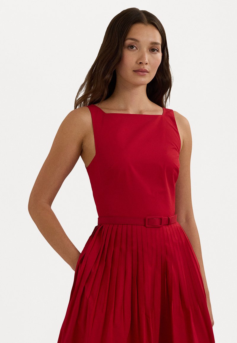 Rochie roșie fără mâneci, cu decolteu pătrat, fustă plisată și o curea asortată. Textură de stofă netedă, design atent croit.