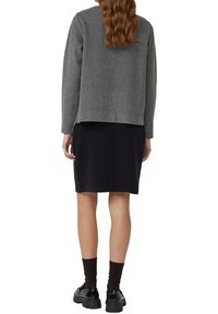 Grijze trui over een zwarte rok, met een losse pasvorm, zachte textuur en een subtiel naaddetail aan de achterkant. Zwarte schoenen en donkere sokken maken de look compleet.