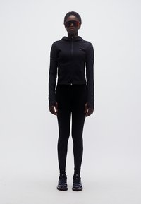 Άτομο που στέκεται φορώντας αθλητική στολή όλη μαύρη, μαύρα παπούτσια τρεξίματος και μεγάλα ανακλαστικά γυαλιά ηλίου, μπροστά σε μια απλή λευκή φόντο.