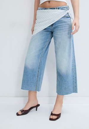 Kobieta w szerokich, skróconych niebieskich dżinsach, z białą, paskowaną tkaniną owiniętą wokół talii oraz ciemnych sandałach na obcasie z odkrytymi palcami.