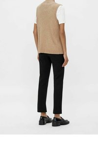 Personne portant un gilet sans manches beige en tricot sur une chemise blanche à manches courtes, un pantalon noir droit et des mocassins noirs à talons, debout devant un fond blanc.