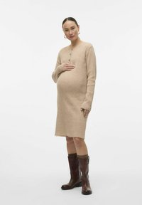 Robe de maternité en tricot, texture côtelée, couleur beige, manches longues, détail de bouton au col. Portée avec des bottes marron à hauteur de genoux.
