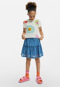 Biela tričko s krátkym rukávom s farebnými kvetinovými a srdcovými aplikáciami, skombinované s modrou džínsovou sukňou s viacerými vrstvami a vyšívanými detailmi. Rúžové sandále.