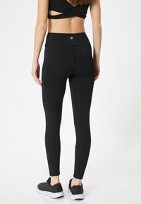 Zwarte high-waisted leggings met een strakke, gladde textuur en subtiele stiksels. De tailleband heeft een klein logo. G gedragen met zwarte sportschoenen.