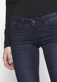 Jeans de denim azul oscuro con un ajuste slim, ligero estiramiento, diseño de cinco bolsillos y cierre frontal con botón. Detalles sutiles de desgaste y desvanecimiento.