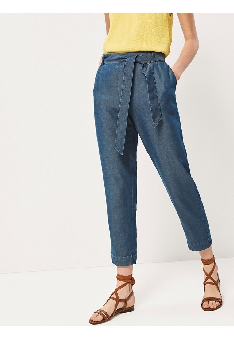 Femme portant un pantalon court bleu taille haute avec une ceinture en tissu, un haut jaune sans manches et des sandales plates marron à brides.